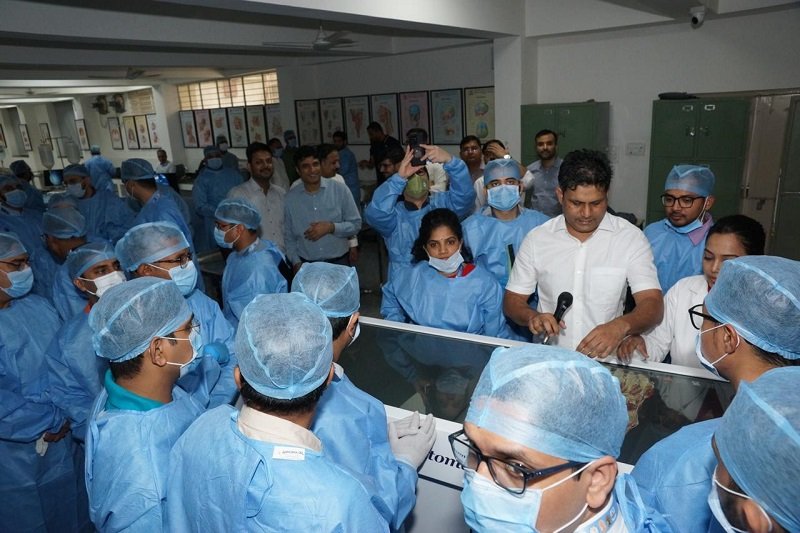 Ishwar Meena guiding a group of medical students during an anatomy lab session, with participants in blue surgical attire focusing on a cadaver dissection.