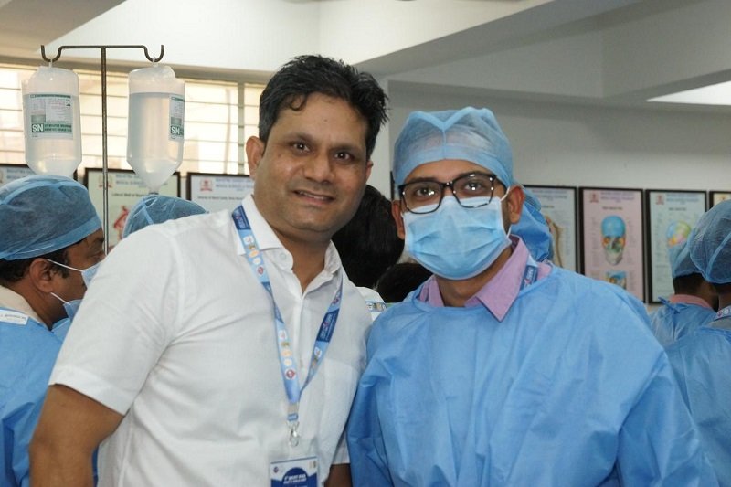 : Ishwar Meena poses with a medical student during an anatomy lab session. The student is dressed in full surgical attire, while Meena, in a white shirt, smiles alongside him, showcasing a collaborative educational environment.