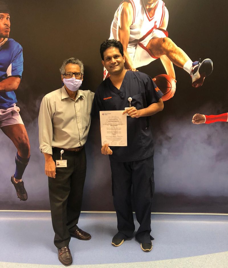 Two men standing side by side; one in a light shirt and black trousers, wearing glasses and a mask, and the other in dark blue scrubs holding a certificate. The background features action shots of athletes in various sports.